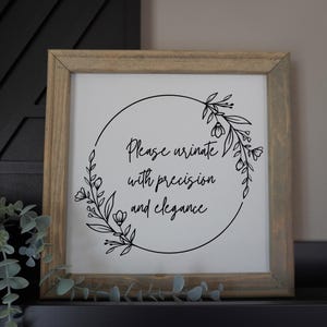 May include: A framed sign with a light wood frame. The white background features a black floral wreath surrounding the text: "Please urinate with precision and elegance."
