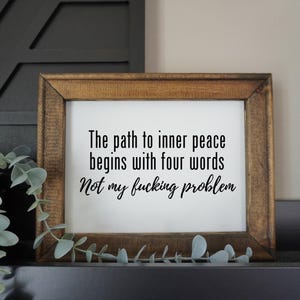 May include: A framed wooden sign with the text "The path to inner peace begins with four words Not my fucking problem" in black. The sign is displayed on a shelf with greenery.