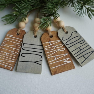May include: Four wooden gift tags with names written in white paint. The tags are tied together with twine and wooden beads. The names on the tags are Josephine, Lucy, Abram, and Sophia.