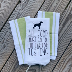 May include: White kitchen towel with green stripes. The towel has a black silhouette of a dog and the text "ALL FOOD MUST GO TO THE LAB FOR TESTING".