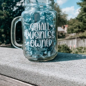 May include: A clear glass mason jar mug with a handle. The mug has the words "SMALL BUSINESS OWNER" written on it in white vinyl lettering. The mug is filled with blue paper shreds.