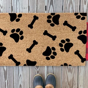 May include: A brown coir doormat with a black paw print and bone pattern. The doormat is on a wooden surface with a red and black plaid rug on either side.
