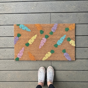 May include: A rectangular doormat with a brown background and a repeating pattern of colourful carrots. The carrots are in shades of blue, yellow, orange, and purple with green tops. White trainers are visible at the bottom of the image.