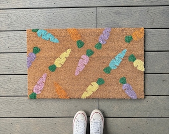 May include: A rectangular doormat with a brown background and a repeating pattern of colorful carrots. The carrots are in shades of blue, yellow, orange, and purple with green tops. White sneakers are visible at the bottom of the image.