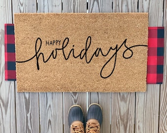 May include: A brown coir doormat with the text "Happy holidays" in black cursive script. The doormat is on a wooden surface and has red and black buffalo plaid fabric on either side.