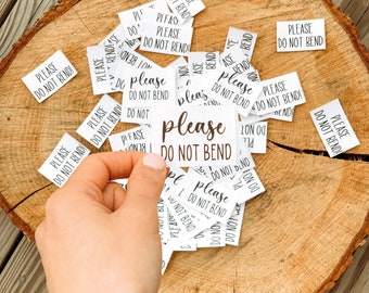 Puede incluir: Un montón de pegatinas de papel blanco con el texto "please do not bend" en ellas. Las pegatinas están esparcidas sobre una superficie de madera.