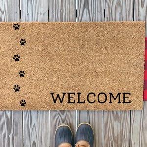 May include: A brown coir doormat with the word "WELCOME" in black lettering. The doormat has a row of black paw prints down the left side. The doormat is framed by red and black buffalo plaid fabric.