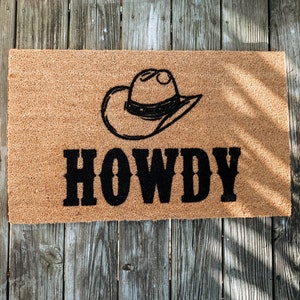 Puede incluir: Una alfombra rectangular con un sombrero de vaquero negro y la palabra "HOWDY" en letras negras en negrita. La alfombra es de color marr&oacute;n natural, hecha de un material &aacute;spero, y descansa sobre una superficie de madera envejecida.