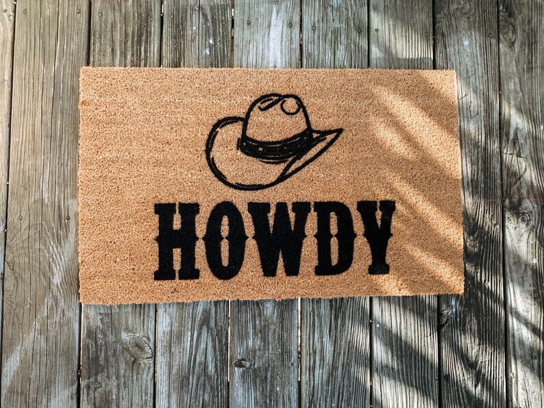 Puede incluir: Una alfombra rectangular con un sombrero de vaquero negro y la palabra "HOWDY" en letras negras en negrita. La alfombra es de color marr&oacute;n natural, hecha de un material &aacute;spero, y descansa sobre una superficie de madera envejecida.