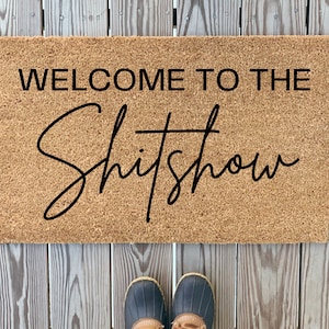 May include: A brown coir doormat with the text "Welcome to the Shitshow" in black lettering.