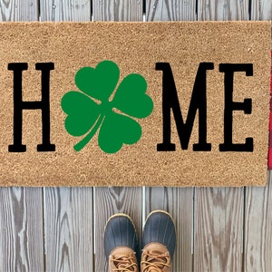 May include: A brown coir doormat with the word "HOME" in black lettering and a green four-leaf clover in the centre.