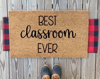 May include: A brown coir doormat with the text "BEST classroom EVER" in black lettering. The doormat is framed by red and black buffalo check fabric.