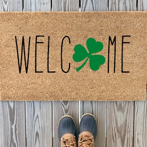 May include: A brown coir doormat with the word "WELCOME" in black lettering and a green shamrock design.
