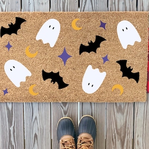 Puede incluir: Una alfombra de coco marrón con temática de Halloween. La alfombra presenta fantasmas blancos, murciélagos negros, medias lunas amarillas y estrellas moradas.