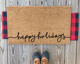 May include: A brown coir doormat with the text "happy holidays" written in black cursive script. The doormat is on a wooden floor and has red and black buffalo plaid fabric on either side.