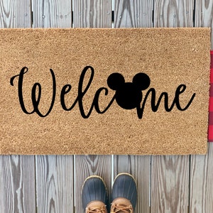 May include: A brown coir doormat with the word "Welcome" in black cursive lettering and a black silhouette of Mickey Mouse's head.