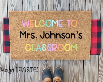 May include: A brown coir doormat with the text "WELCOME TO Mrs. Johnson's CLASSROOM" in pastel colored letters.