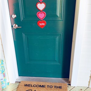 May include: A green door with a Valentine's Day themed door hanging featuring red and pink hearts with white polka dots. The door hanging says "Welcome" and "XOXO". A welcome mat with the text "Welcome to the Shitshow" is on the floor in front of the door.