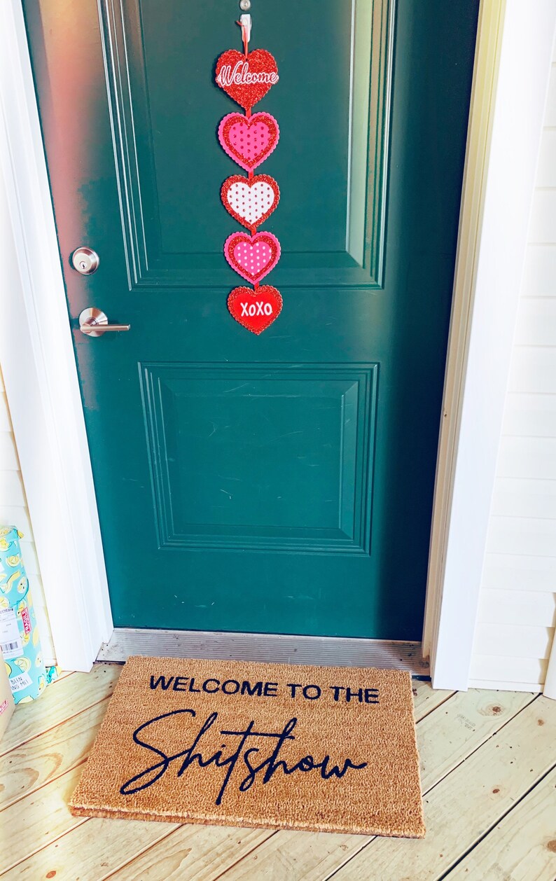 May include: A green door with a Valentine's Day themed door hanging featuring red and pink hearts with white polka dots. The door hanging says "Welcome" and "XOXO". A welcome mat with the text "Welcome to the Shitshow" is on the floor in front of the door.