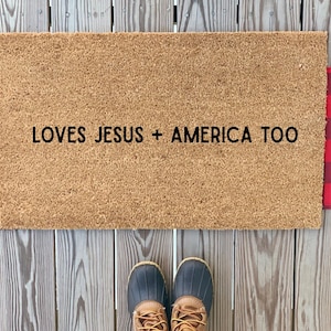 May include: A rectangular doormat with the text "LOVES JESUS + AMERICA TOO" in black. The mat is bordered with red and black buffalo plaid fabric. A pair of boots is visible at the bottom.