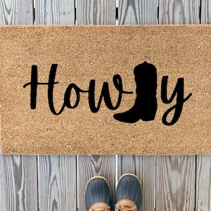 May include: A brown coir doormat with the text "Howdy" in black cursive lettering and a black silhouette of a cowboy boot.