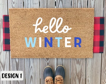 Op de afbeelding: Een rechthoekige deurmat met de woorden "hello WINTER" in witte en blauwe letters. De mat heeft een natuurlijk bruin kokosoppervlak en wordt geflankeerd door rood en zwart geblokte buffelstof. Het ontwerp is gelabeld "DESIGN 1".