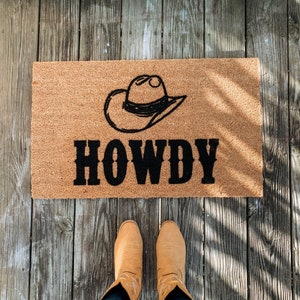 Puede incluir: Una alfombra rectangular hecha de fibras naturales con la palabra "HOWDY" en letras negras en negrita y un gr&aacute;fico de sombrero de vaquero negro. La alfombra est&aacute; sobre un porche de madera, con un par de botas de vaquero marrones y vaqueros azules.