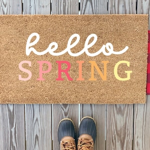 May include: A brown coir doormat with the text "hello spring" in white, pink, and orange lettering. The doormat is on a wooden surface with a red and black plaid rug on either side.