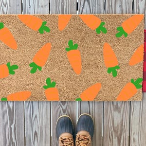 Könnte beinhalten: Eine rechteckige Fußmatte mit einem Hintergrund aus Naturfaser, verziert mit orangefarbenen Karotten und grünen Spitzen. Die Matte wird von rot-schwarz kariertem Stoff flankiert. Ein Paar blaue und beige Stiefel befindet sich am unteren Bildrand.