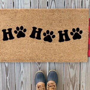 May include: A rectangular doormat with a light brown surface, featuring the text "H🐾H🐾H🐾" in black. The mat has red and black plaid accents on each side. A pair of boots is visible at the bottom.