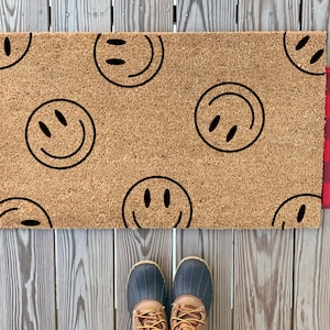 May include: A brown coir doormat with a black and white checkered border. The doormat has a repeating pattern of black smiley faces.