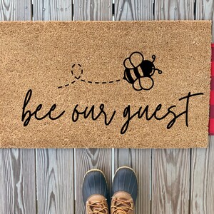 May include: A brown coir doormat with the text "bee our guest" in black cursive font. A black and yellow cartoon bee with wings is flying above the text. The doormat is on a wooden surface.