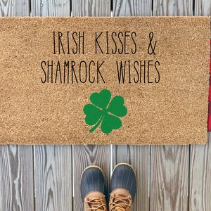 May include: A brown coir doormat with the text "Irish Kisses & Shamrock Wishes" in black lettering. A green shamrock is centered on the mat. The mat is flanked by red and black buffalo plaid fabric.