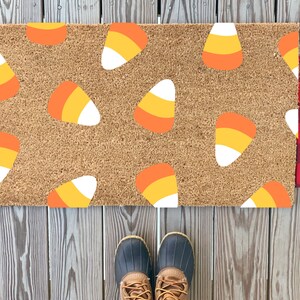 Puede incluir: Una alfombra de coco marrón con un tema de Halloween que presenta un patrón repetido de maíz dulce naranja, amarillo y blanco.