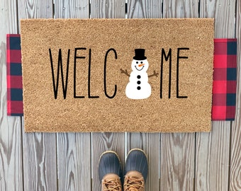 May include: A brown coir doormat with the word "WELCOME" in black lettering and a snowman graphic in the center. The doormat is on a wooden surface.
