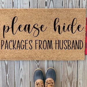 May include: A brown coir doormat with the text "please hide packages from husband" in black lettering. The doormat is surrounded by a red and black plaid fabric.