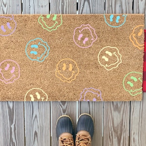 May include: A brown coir doormat with a variety of colorful smiley faces printed on it. The doormat is placed on a wooden floor.