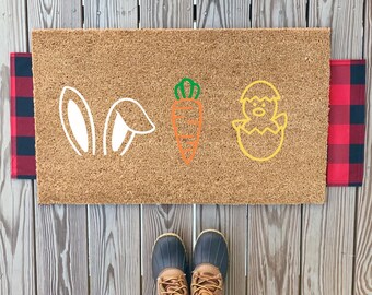 May include: A brown coir doormat with white bunny ears, an orange carrot, and a yellow chick in an egg. The doormat is on a wooden deck.
