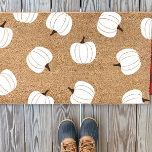 May include: A brown coir doormat with a white pumpkin pattern. The doormat is on a wooden deck with red and black plaid fabric on either side.