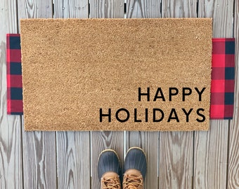 May include: A brown coir doormat with the text "HAPPY HOLIDAYS" in black lettering. The doormat is on a wooden surface with red and black buffalo plaid fabric on either side.