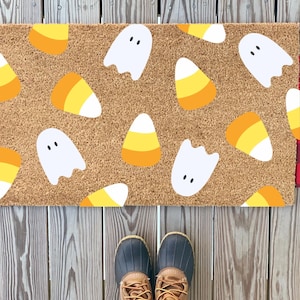 May include: A rectangular doormat with a brown background features a pattern of white ghosts and candy corn. The sides of the mat have red and black plaid fabric. The mat is placed on a wooden deck.