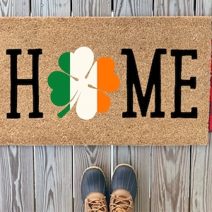 Puede incluir: Una alfombra de coco marrón con la palabra "HOME" en letras negras y un trébol verde, blanco y naranja en el centro.