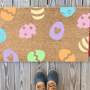 May include: A brown coir doormat with a colorful Easter egg design. The eggs are in various shades of pink, blue, green, and yellow. The doormat is on a wooden deck.