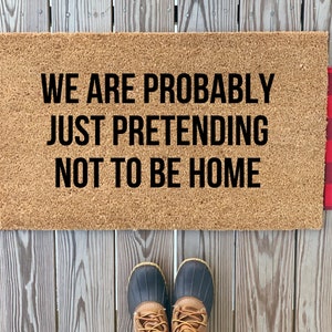 以下が含まれることがあります： 「WE ARE PROBABLY JUST PRETENDING NOT TO BE HOME」と黒字で書かれた長方形のドアマット。 マットは自然な茶色で、両側に赤と黒のバッファローチェックのアクセントがあります。 下にはブーツが1足あります。