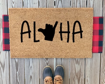May include: A brown coir doormat with the word "ALOHA" in black lettering and a black hand gesture. The doormat is on a wooden surface with red and black checkered fabric on either side.