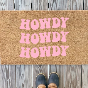 Może przedstawiać: Brązowa mata kokosowa z napisem "HOWDY" powtórzonym trzy razy w różowych, retro literach.