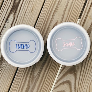 May include: Two white dog bowls with bone-shaped name tags. The bowl on the left has a blue bone with the name "TUCKER" and the bowl on the right has a pink bone with the name "SADIE".