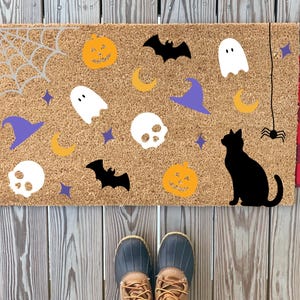 May include: A Halloween-themed doormat with a natural coir background. It features orange pumpkins, white ghosts, black bats, purple witch hats, and a black cat silhouette. A spider web and spider are also present.