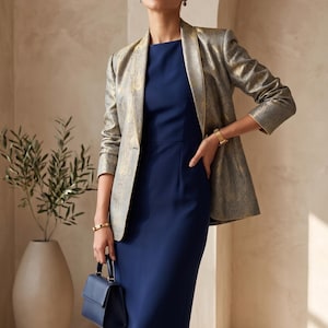 May include: A navy blue midi dress with a round neckline paired with a metallic gold and grey patterned blazer featuring long sleeves and a structured lapel. A small navy blue handbag with a top handle is held in one hand. The text Nibs tog is visible.
