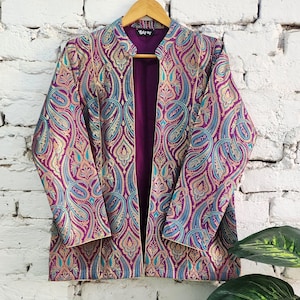 May include: A colourful open-front jacket featuring a paisley design in purple, teal, and gold. The jacket has a deep purple lining and is suspended on a wooden hanger against a white brick wall. The intricate design of the jacket is eye-catching.